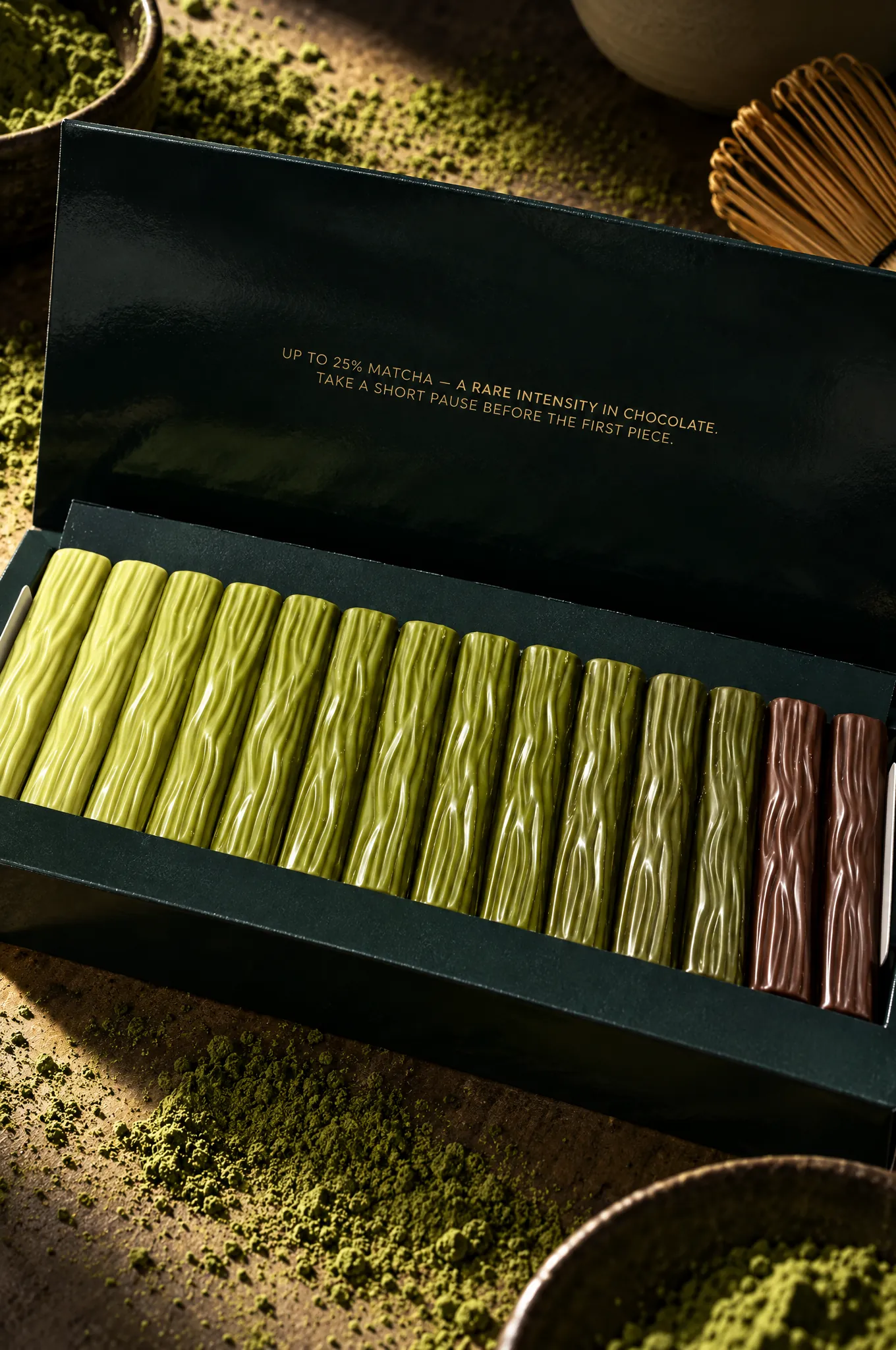 Open box of 7 Shades of Matcha Chocolate with sticks from light to dark green, matcha powder and a bamboo whisk